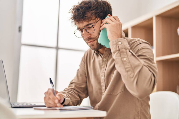Jovem trabalhador de negócios falando no smartphone escrevendo no notebook no escritório - Foto, Imagem