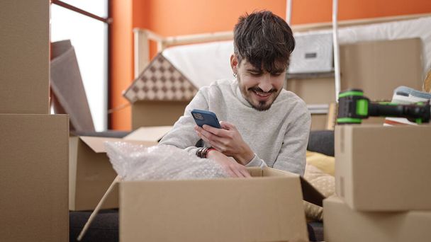 Молодой латиноамериканец распаковывает картонную коробку со смартфона в новом доме - Фото, изображение