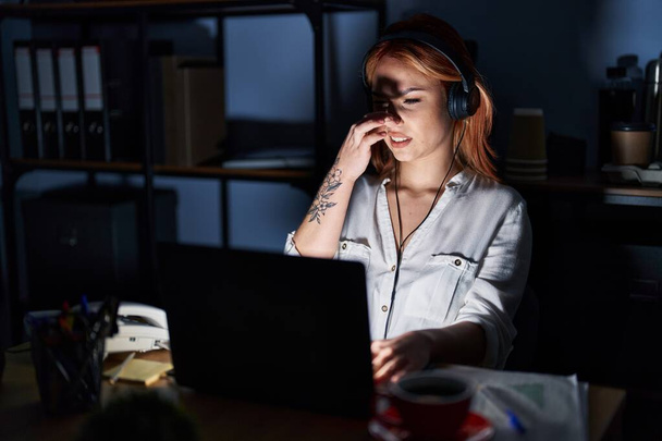 Young caucasian woman working at the office at night smelling something stinky and disgusting, intolerable smell, holding breath with fingers on nose. bad smell  - Foto, afbeelding