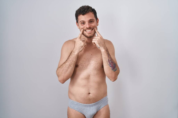 Fiatal spanyol férfi áll félmeztelenül fehérneműben mosolyogva nyitott szájjal, ujjal mutogatva és vidám mosolyt erőltetve  - Fotó, kép