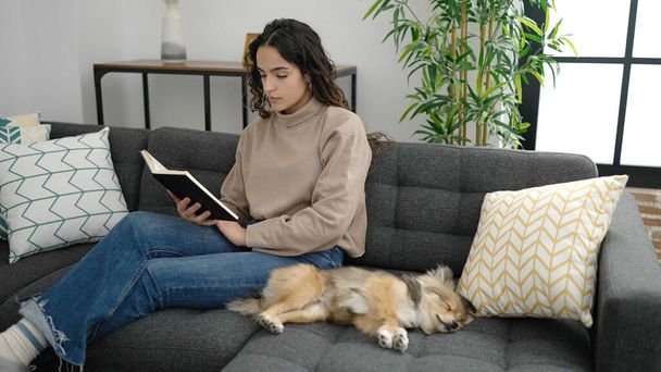 Young hispanic woman with dog reading book sitting on sofa at home - Фото, изображение