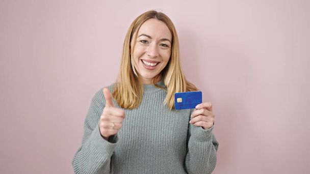 Junge blonde Frau lächelt selbstbewusst und zeigt auf Kreditkarte vor isoliertem rosa Hintergrund - Foto, Bild