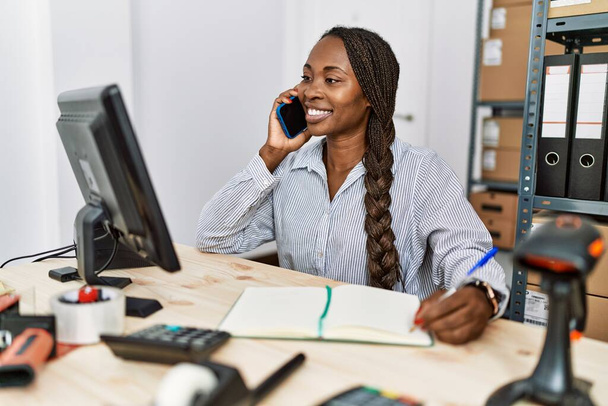 Mujer afroamericana comercio electrónico trabajador de negocios hablando en la escritura de teléfonos inteligentes en el cuaderno en la oficina - Foto, imagen