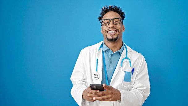 Afro-américain médecin souriant en utilisant smartphone sur fond bleu isolé - Photo, image