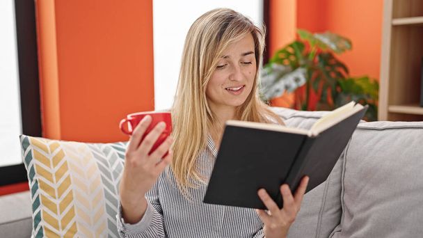 Jeune femme blonde lecture livre et boire du café assis sur le canapé à la maison - Photo, image