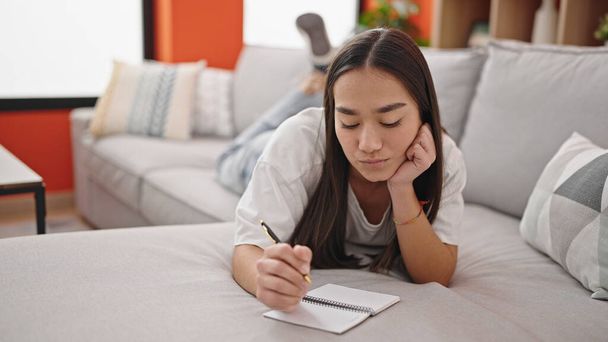 Młoda, piękna latynoska kobieta pisząca na notatniku leżąca na sofie w domu - Zdjęcie, obraz