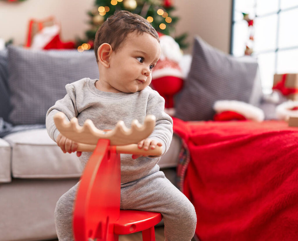 Adorable hispanic baby playing with reindeer rocking by christmas tree at home - Фото, зображення