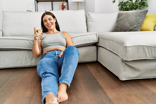 Молода красива іспаномовна жінка п'є келих вина, сидячи на підлозі вдома
 - Фото, зображення