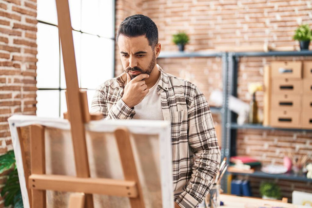 Giovane uomo ispanico guardando disegnare con espressione dubbio a studio d'arte - Foto, immagini