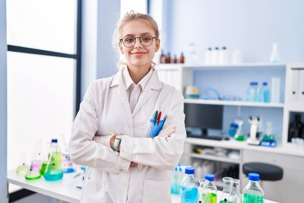 Молодая блондинка ученый улыбается уверенно стоя со скрещенными руками в лаборатории - Фото, изображение