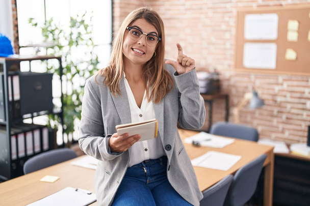 Junge hispanische Frau, die im Büro arbeitet, trägt eine Brille, lächelt und gestikuliert selbstbewusst mit der Hand und macht ein kleines Zeichen mit den Fingern und der Kamera. Maßnahmenkonzept.  - Foto, Bild