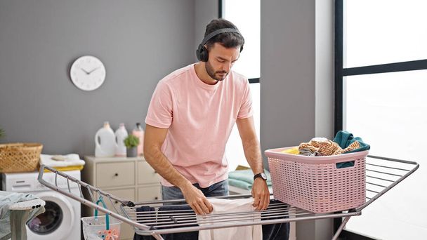 Young hispanic man listening to music hanging clothes on clothesline at laundry room - Foto, imagen