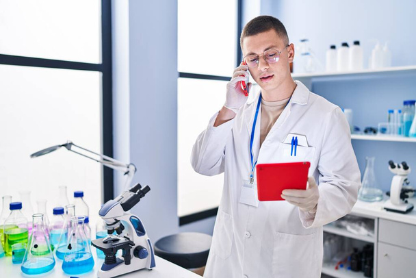 Young hispanic man scientist talking on smartphone using touchpad at laboratory - Photo, Image