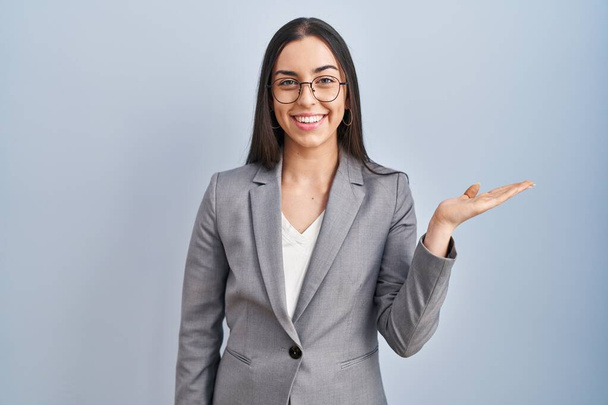 Hispanic business woman wearing glasses smiling cheerful presenting and pointing with palm of hand looking at the camera.  - Foto, Imagen