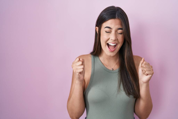 Femme hispanique debout sur fond rose excité pour le succès avec les bras levés et les yeux fermés célébrant la victoire en souriant. concept gagnant.  - Photo, image