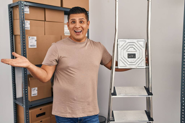 Hispanic young man working at small business ecommerce holding ladders celebrating achievement with happy smile and winner expression with raised hand  - Zdjęcie, obraz