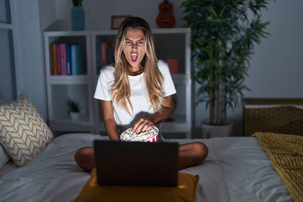 Jonge blonde vrouw zit thuis op het bed naar een film te kijken vanaf een laptop boos en boos schreeuwend gefrustreerd en woedend, schreeuwend van woede. woede en agressief concept.  - Foto, afbeelding