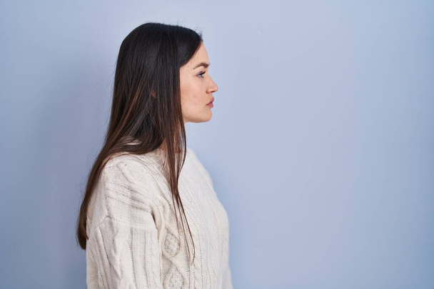 Jeune femme brune debout sur fond bleu regardant vers le côté, pose de profil relax avec visage naturel et sourire confiant.  - Photo, image