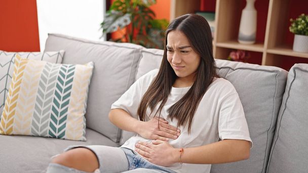 Νεαρή όμορφη ισπανόφωνη γυναίκα που υποφέρει για πόνο στο στομάχι κάθεται στον καναπέ στο σπίτι - Φωτογραφία, εικόνα