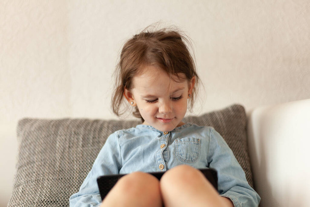 Nettes kleines Mädchen sitzt auf dem Sofa und spielt zu Hause mit dem Smartphone - Foto, Bild