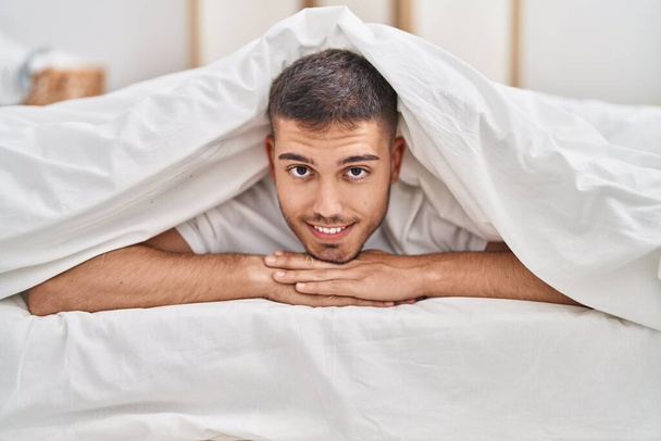 Jeune homme hispanique couvrant avec drap couché sur le lit dans la chambre - Photo, image