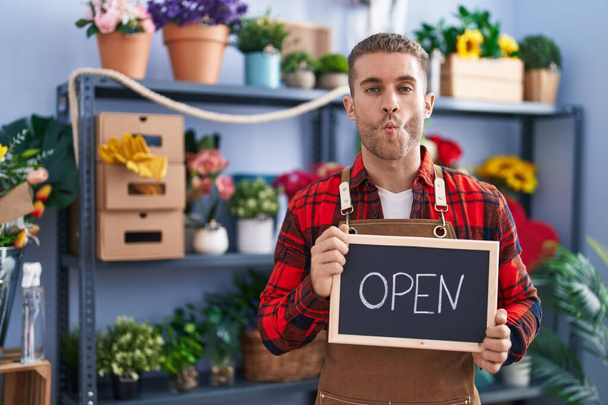 Young caucasian man working at florist holding open sign making fish face with mouth and squinting eyes, crazy and comical.  - Photo, Image