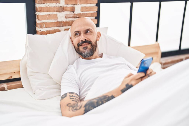 Young bald man using smartphone lying on bed at bedroom - Фото, изображение