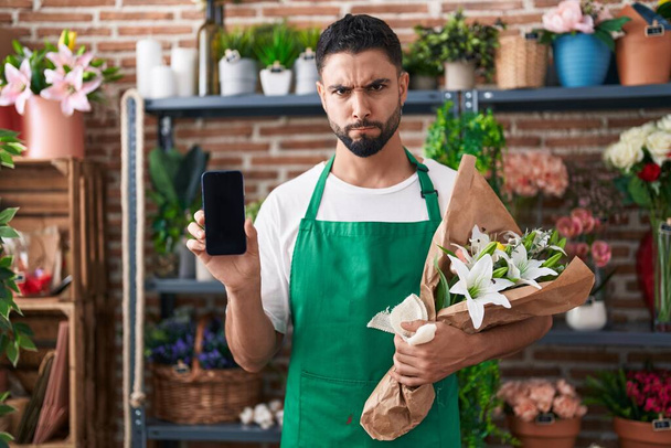 Joven hispano que trabaja en floristería mostrando escéptico y nervioso de la pantalla del teléfono inteligente, frunciendo el ceño molesto debido a un problema. persona negativa.  - Foto, imagen