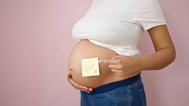 Young pregnant woman pointing to girl reminder paper on belly over isolated pink background - Fotoğraf, Görsel