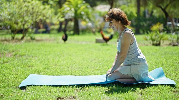Middelbare leeftijd vrouw zittend op knieën op yoga mat met serieus gezicht in park - Foto, afbeelding