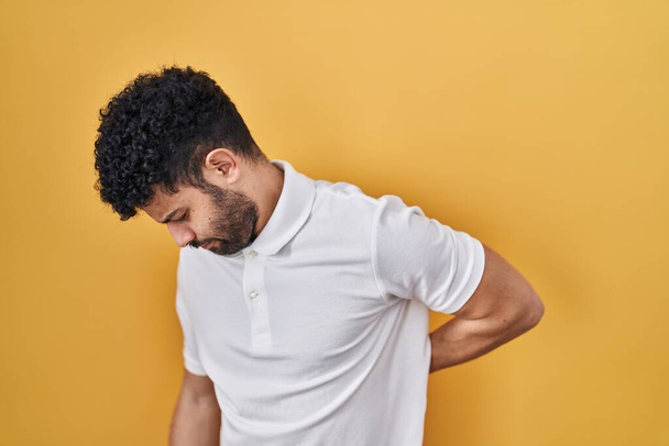 Arabský muž stojící nad žlutým pozadím trpící bolestí zad, dotýkající se rukou, svalová bolest  - Fotografie, Obrázek