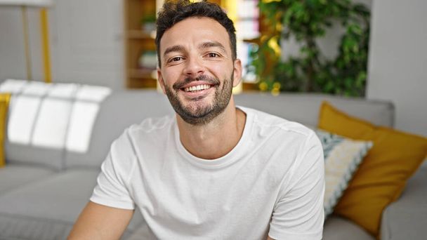Jonge Spaanse man die thuis op de bank zit te glimlachen - Foto, afbeelding