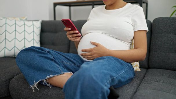 Młoda kobieta w ciąży za pomocą smartfona siedzi na kanapie w domu - Zdjęcie, obraz
