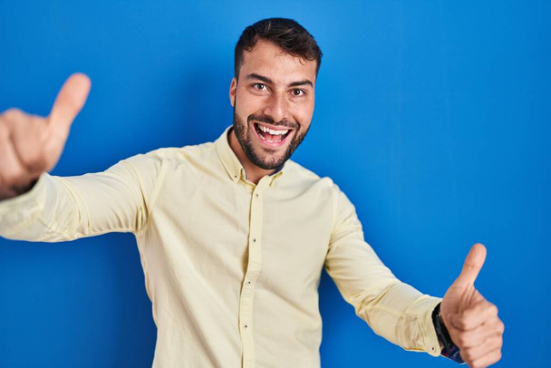 青い背景の上に立つハンサムなヒスパニック系の男性は、手で積極的なジェスチャーを行うことを承認し、親指を上げて笑顔と成功のために幸せ。勝者のジェスチャー.  - 写真・画像