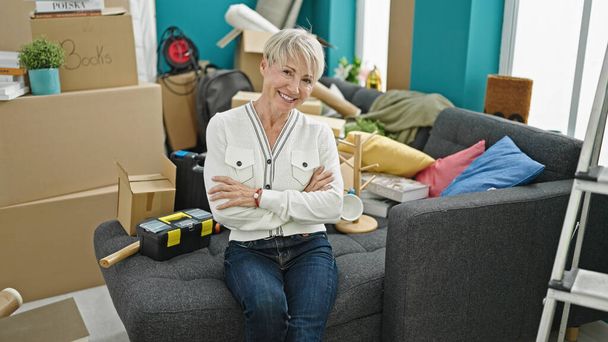 Middle age blonde woman smiling confident sitting with arms crossed gesture at new home - Photo, Image