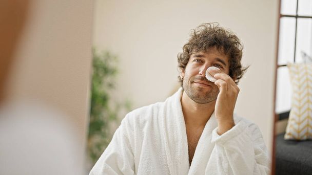 Νεαρός Ισπανός άνδρας που φοράει μπουρνούζι καθαρίζοντας το πρόσωπο με βαμβακερό μαξιλάρι κοιτώντας στον καθρέφτη στο σπίτι - Φωτογραφία, εικόνα