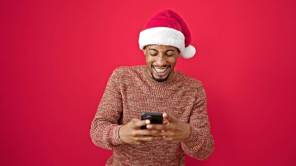アフリカ系アメリカ人男性身に着けているクリスマスの帽子使用スマートフォン笑顔上の隔離された赤の背景 - 写真・画像