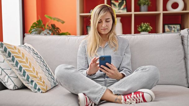 Young blonde woman listening to music sitting on sofa at home - 写真・画像