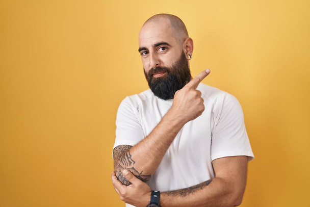Young hispanic man with beard and tattoos standing over yellow background pointing with hand finger to the side showing advertisement, serious and calm face  - Foto, Imagen