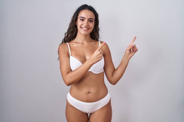 Junge hispanische Frau in weißer Dessous lächelt und schaut in die Kamera, die mit zwei Händen und Fingern zur Seite zeigt.  - Foto, Bild