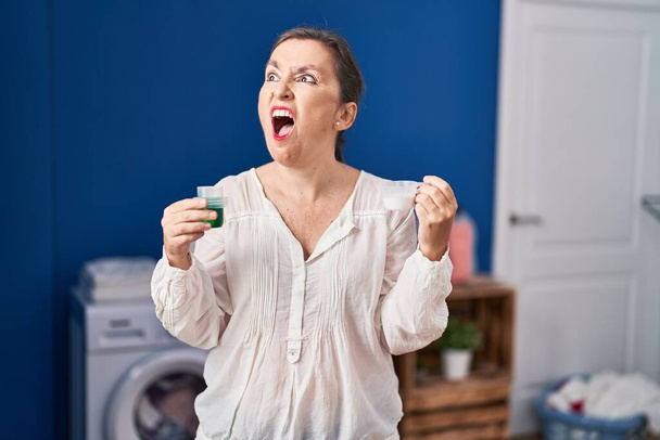 Μεσήλικας Ισπανίδα που κρατάει απορρυπαντικό ρούχων και σκόνη πλυντηρίου θυμωμένη και θυμωμένη ουρλιάζοντας απογοητευμένη και έξαλλη, φωνάζοντας με θυμό. οργή και επιθετική αντίληψη.  - Φωτογραφία, εικόνα