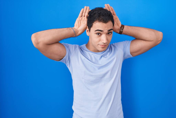 Young hispanic man standing over blue background doing bunny ears gesture with hands palms looking cynical and skeptical. easter rabbit concept.  - Foto, immagini