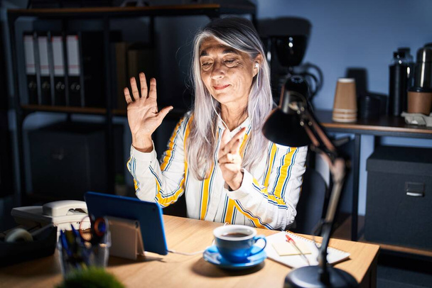 Жінка середнього віку з сірим волоссям працює в офісі вночі показуючи і вказуючи пальцями номер вісім, посміхаючись впевнено і щасливо.  - Фото, зображення