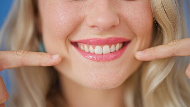 Young blonde woman pointing to teeth with fingers over isolated blue background - 写真・画像