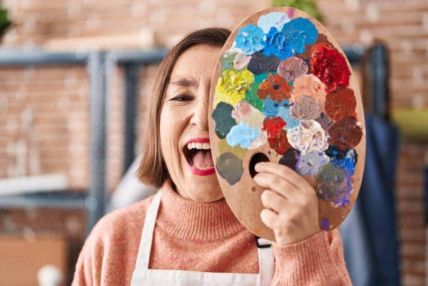 Middle age hispanic woman holding painter palette close to face smiling and laughing hard out loud because funny crazy joke.  - Fotografie, Obrázek