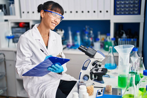 Young beautiful hispanic woman scientist smiling confident writing on document at laboratory - Photo, Image