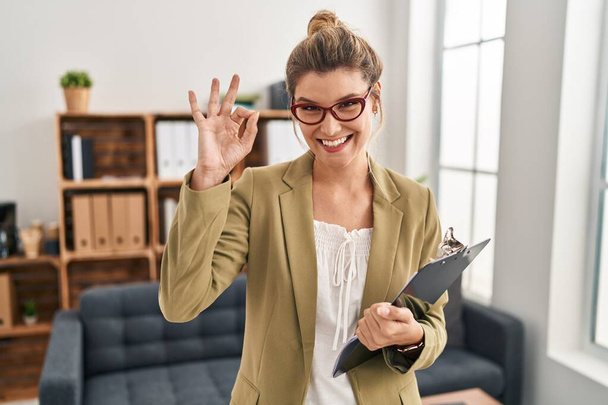 Молодая женщина, работающая в консультационном офисе улыбаясь позитивно делает хорошо знак рукой и пальцами. успешное выражение.  - Фото, изображение