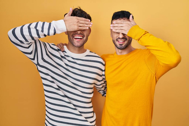Молодая латиноамериканская гей-пара, стоящая на жёлтом фоне, улыбаясь и смеясь, закрывая глаза рукой. слепая концепция.  - Фото, изображение