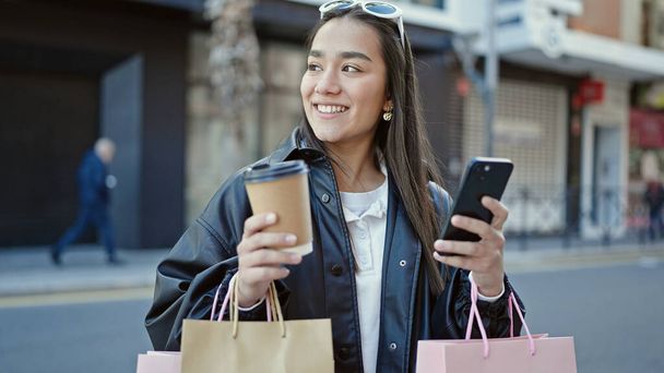 Genç ve güzel İspanyol kadın akıllı telefon kullanıyor. Elinde alışveriş poşetleri ve sokakta kahve tutuyor. - Fotoğraf, Görsel