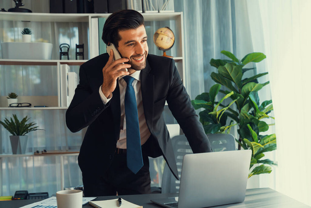 Fleißiger Geschäftsmann, der mit Kunden telefoniert, während er in seinem Büro mit Laptop arbeitet, als Konzept eines modernen, hart arbeitenden Büroarbeiterlebens mit Mobiltelefon. Inbrünstig - Foto, Bild